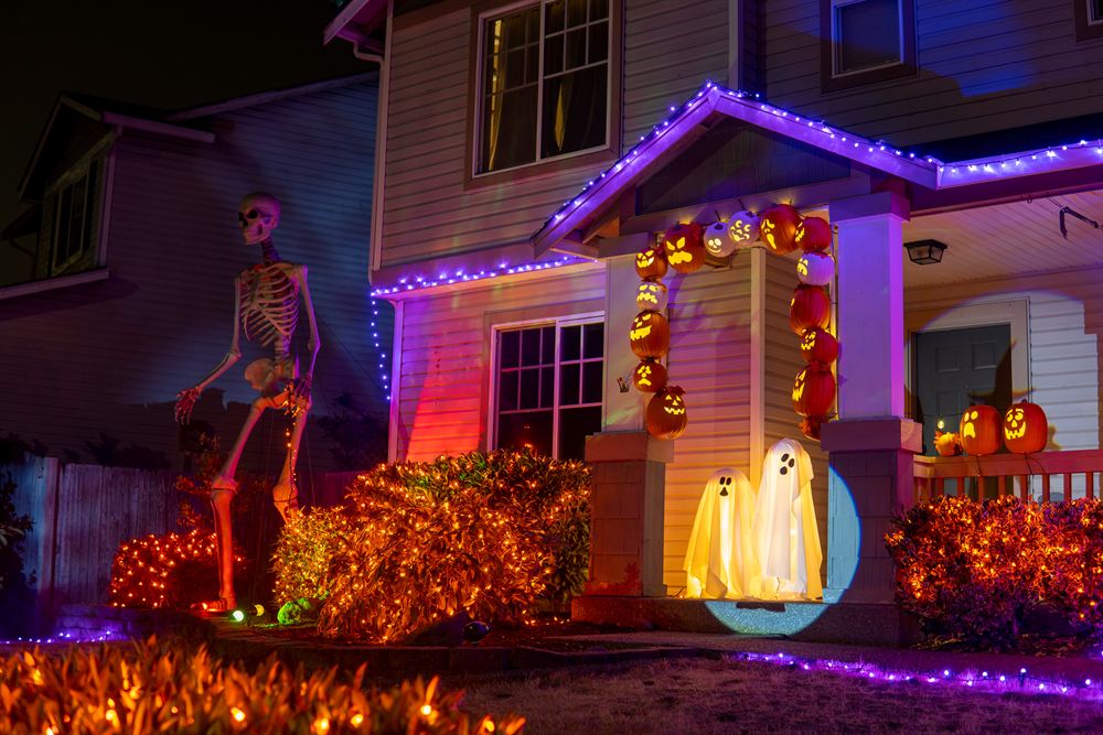 A house decorated for Halloween with purple roofline lights, orange-lit hedges, glowing ghosts and jack-o'-lanterns